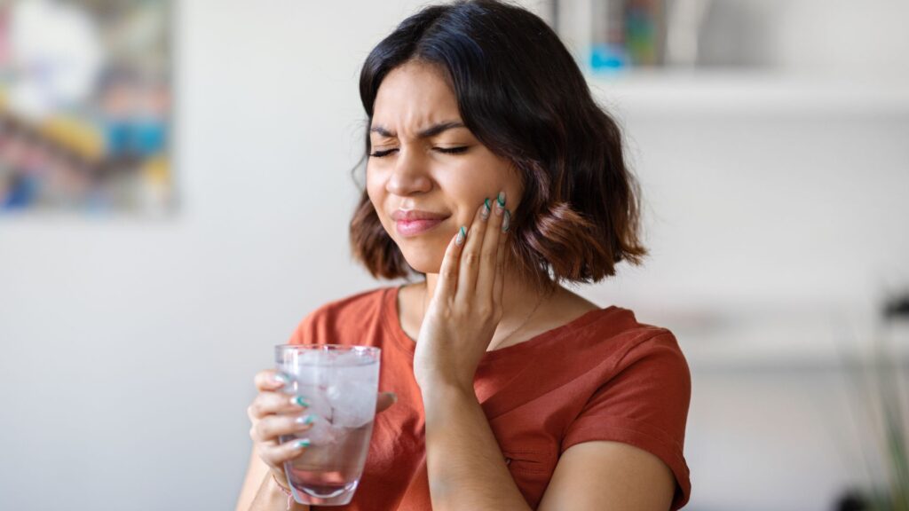 Woman wincing in pain from drinking ice water, showing symptoms of sensitive teeth.